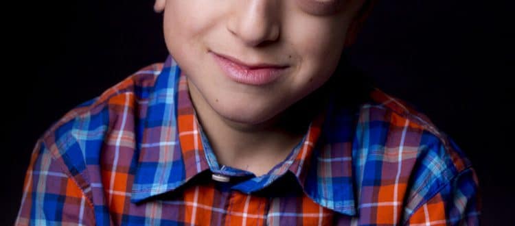 Ritratto ravvicinato di un bambino con sindrome di Crouzon che sorride, indossa una camicia a quadri colorati su sfondo nero – Altravoce Onlus