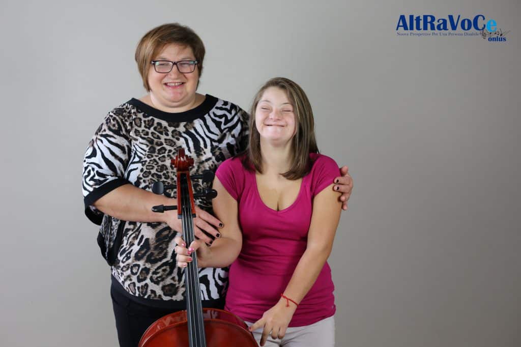 Due donne sorridenti, una adulta e una giovane con sindrome di Down, posano insieme tenendo un violoncello, simbolo di relazione, inclusione e partecipazione musicale.